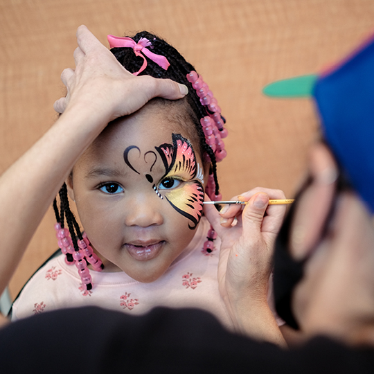 a girl with braids and pink beads is getting a butterfly wing painted on the right side of her face.