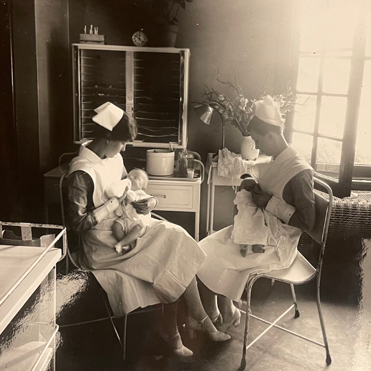 An old black and white photo showing two women in white nursing uniforms hold and feed two babies.