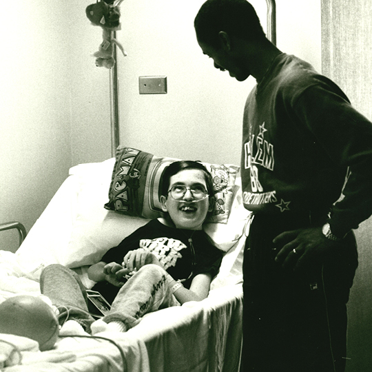 A man stands and talks to a patient who is laying in a bed smiling.