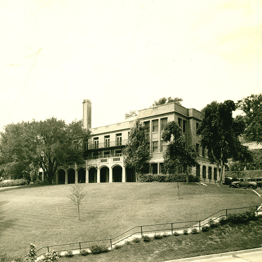 A large brick building sits on top of a hill. To the right are two1930s model cars.