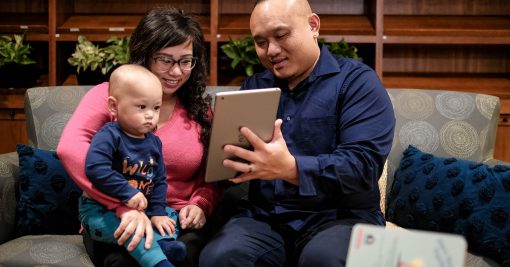 a woman with black hair holds her young son on her lap. next to them, the boy's father is holding up an iPad for them to use. They are all smiling and looking at the iPad.
