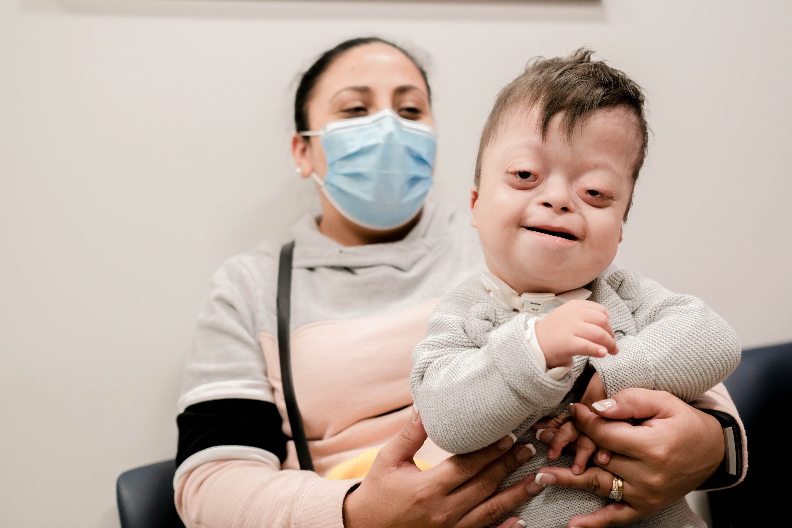 A young boy sits on his mother's lap. She has a medical mask on.