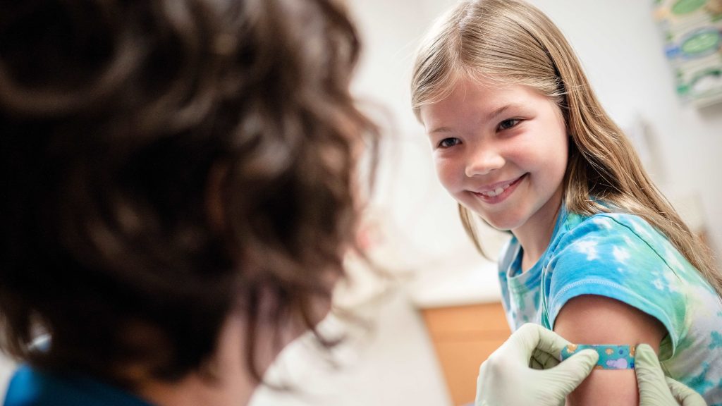 Girl smiling while adult puts a band aid on her arm.