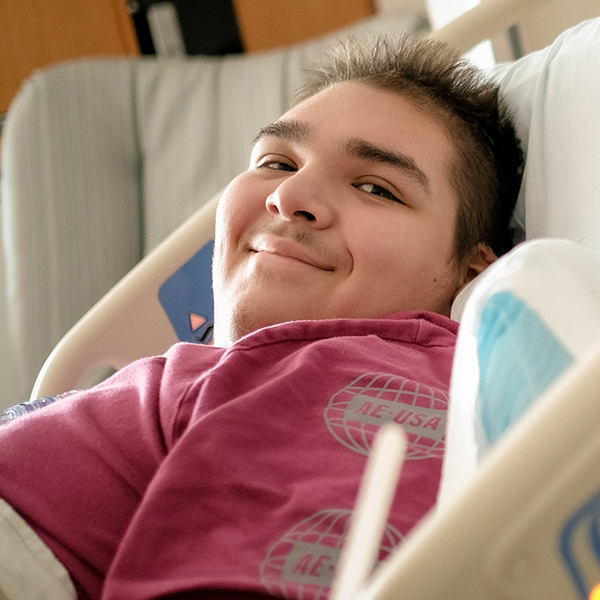 A teenage boy lies in a hospital bed. He's smiling at the camera and wearing a red shirt.