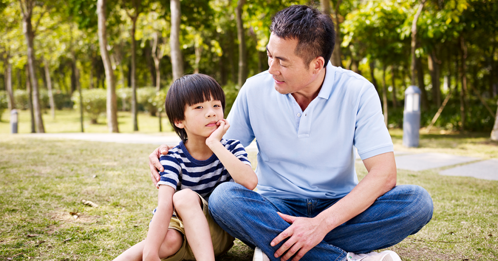 Asian father and son having a conversation outside sitting in the grass