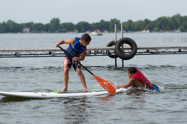 A camper paddles on the paddleboard while another camper tries to swim up