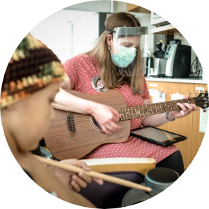 A masked woman plays an acoustic guitar, blending her passion for music therapy and commitment to health precautions.