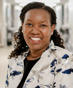 Dr. Caroline Njau is wearing a floral print jacket and is smiling at the camera.
