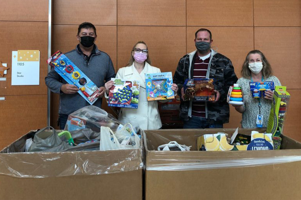 A group of 4 adults stand in front of a wall that says Star Studio. They're all wearing masks and holding toys. In the foreground are two big boxes full of toys.