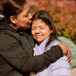 Neli, a teenage girl stands outside with her mom. Neli is smiling with her eyes closed while her mom kisses her on the forhead.