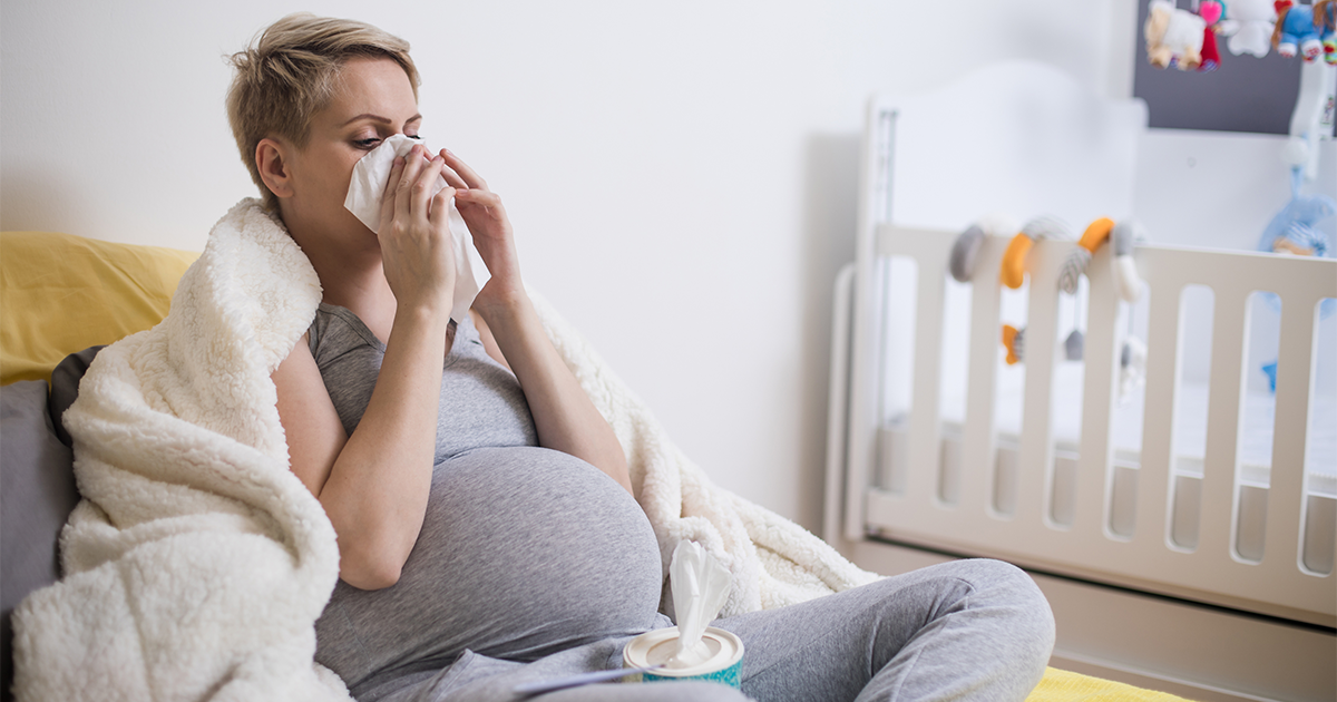 Pregnant woman blowing nose into a tissue while sick at home