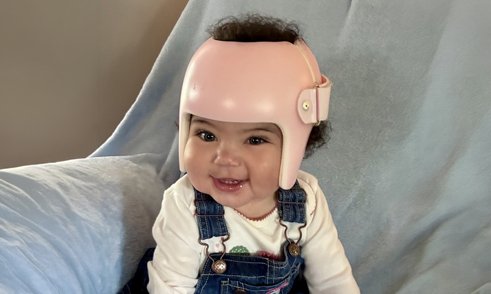 A smiling baby wearing a pink medical helmet, seated on a light blue blanket. The baby is dressed in denim overalls and a white shirt.