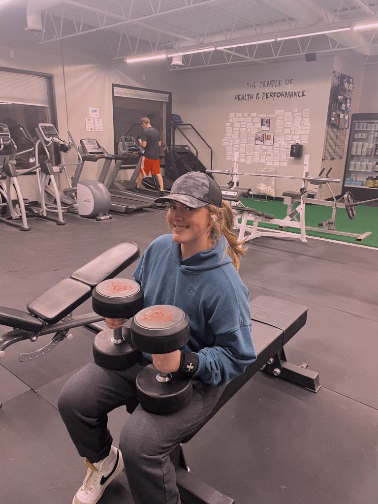 Gwen in a hat at the gym holding two dumbbells