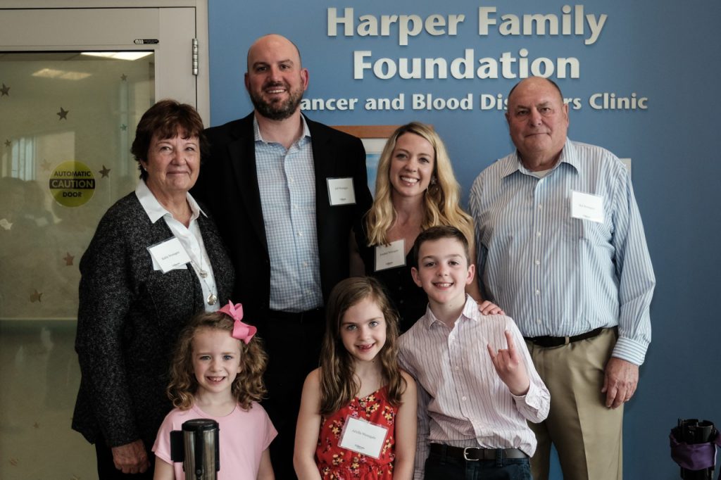 A family stands in front of a sign that says "Harper Family Foundation." There are four adults standing in the back row, and in front are 2 girls and a boy. Everyone is smiling at the camera.
