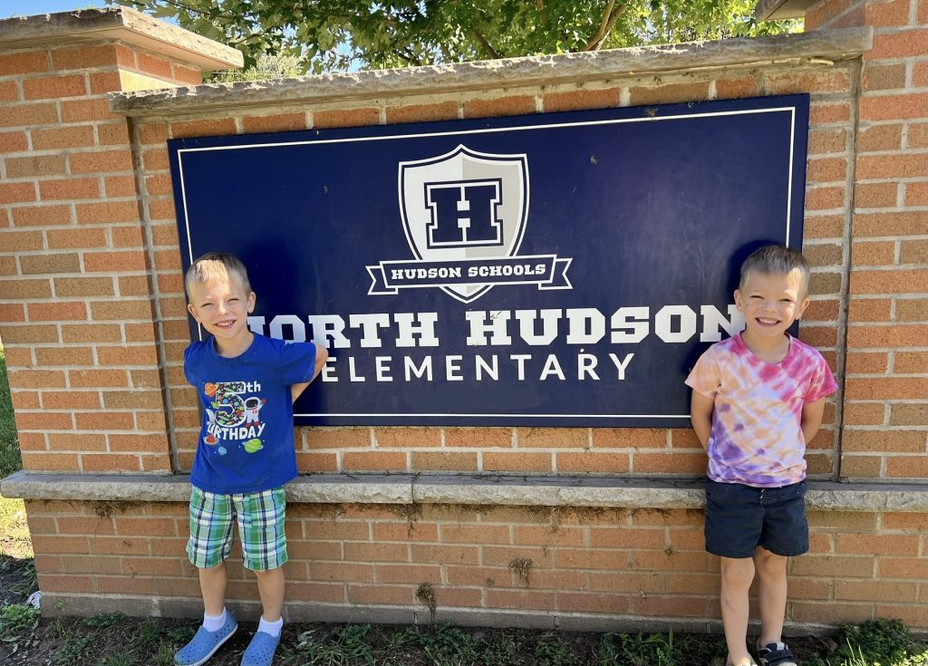 Axel and Barrett enter kindergarten and smile in front of their school's sign
