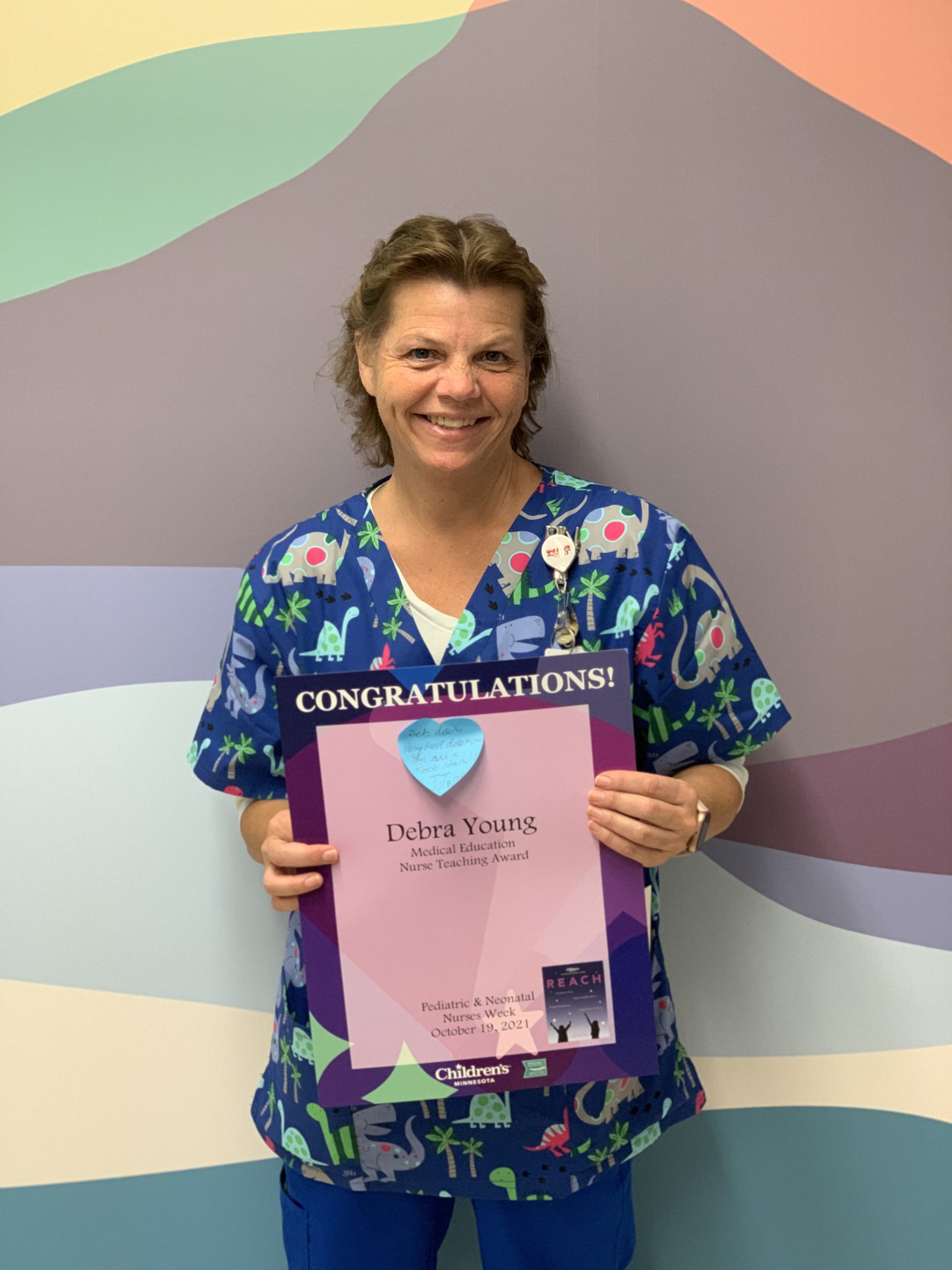 A woman with curly brown hair pulled back, wears dinosaur print scrubs. She's smiling and holding a sign that says Congratulations!