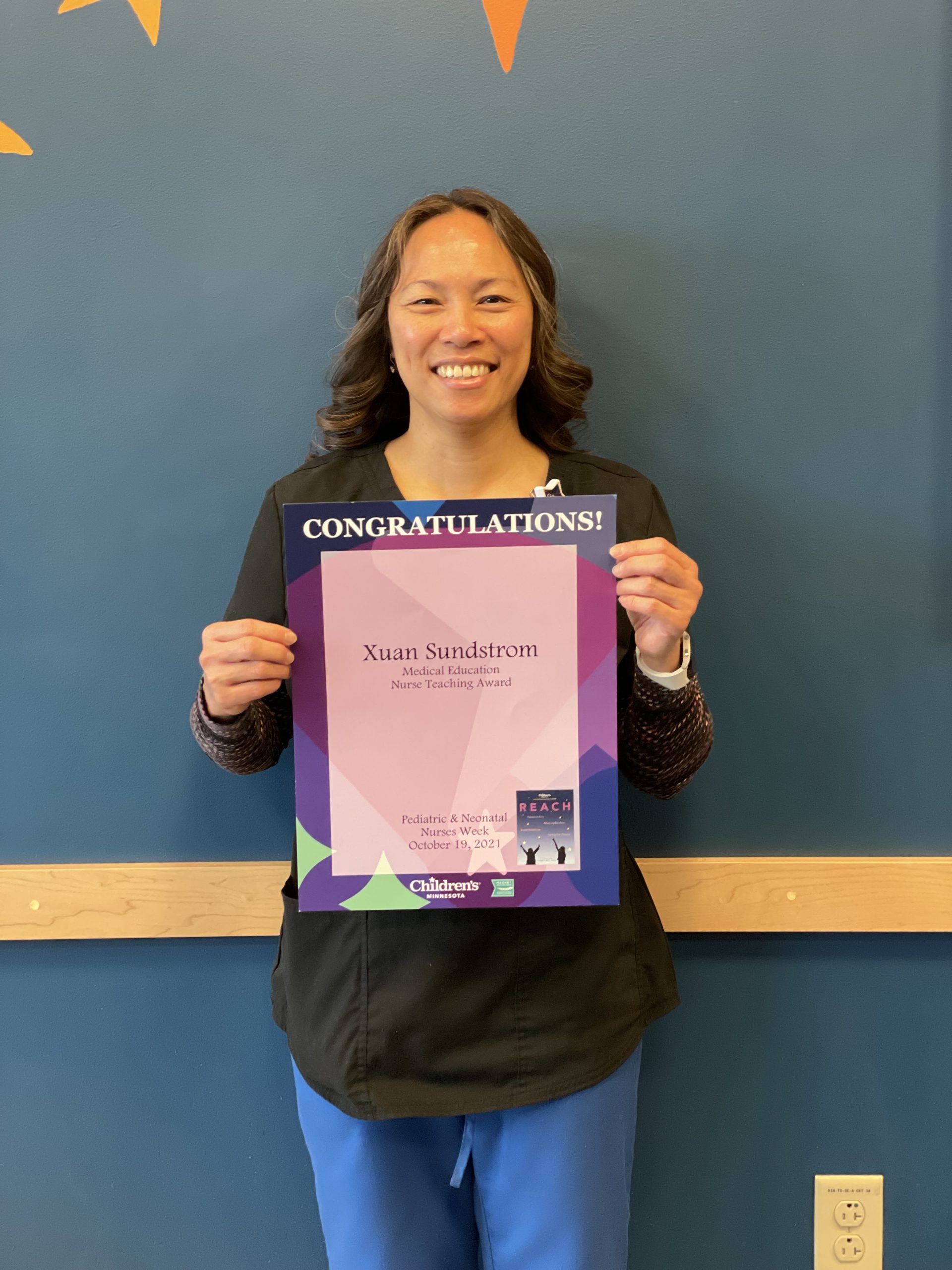 A woman with long dark hair is wearing a black shirt and blue pants. She's smiling and holding up a sign that says Congratulations!