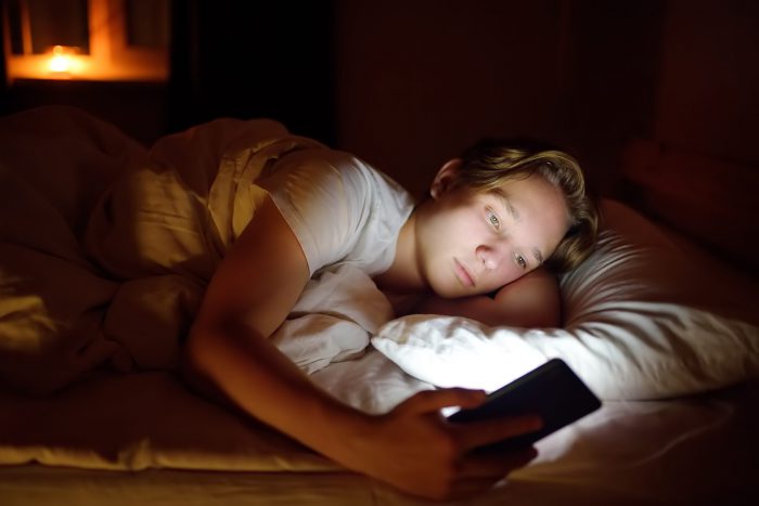 Teenager boy chatting by phone, writing in blog or reading social networks instead sleeping. Young guy laying in bed and watching video clips by smartphone. Overuse and addiction kids from gadgets.