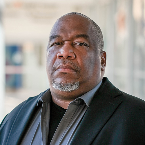 James Burroughs, a black man, is looking at the camera with a serious expression. He is wearing a gray shirt and a black suit jacket.