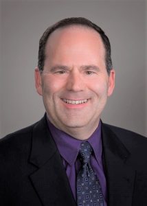 John Harding smiles in purple shirt and black suit.