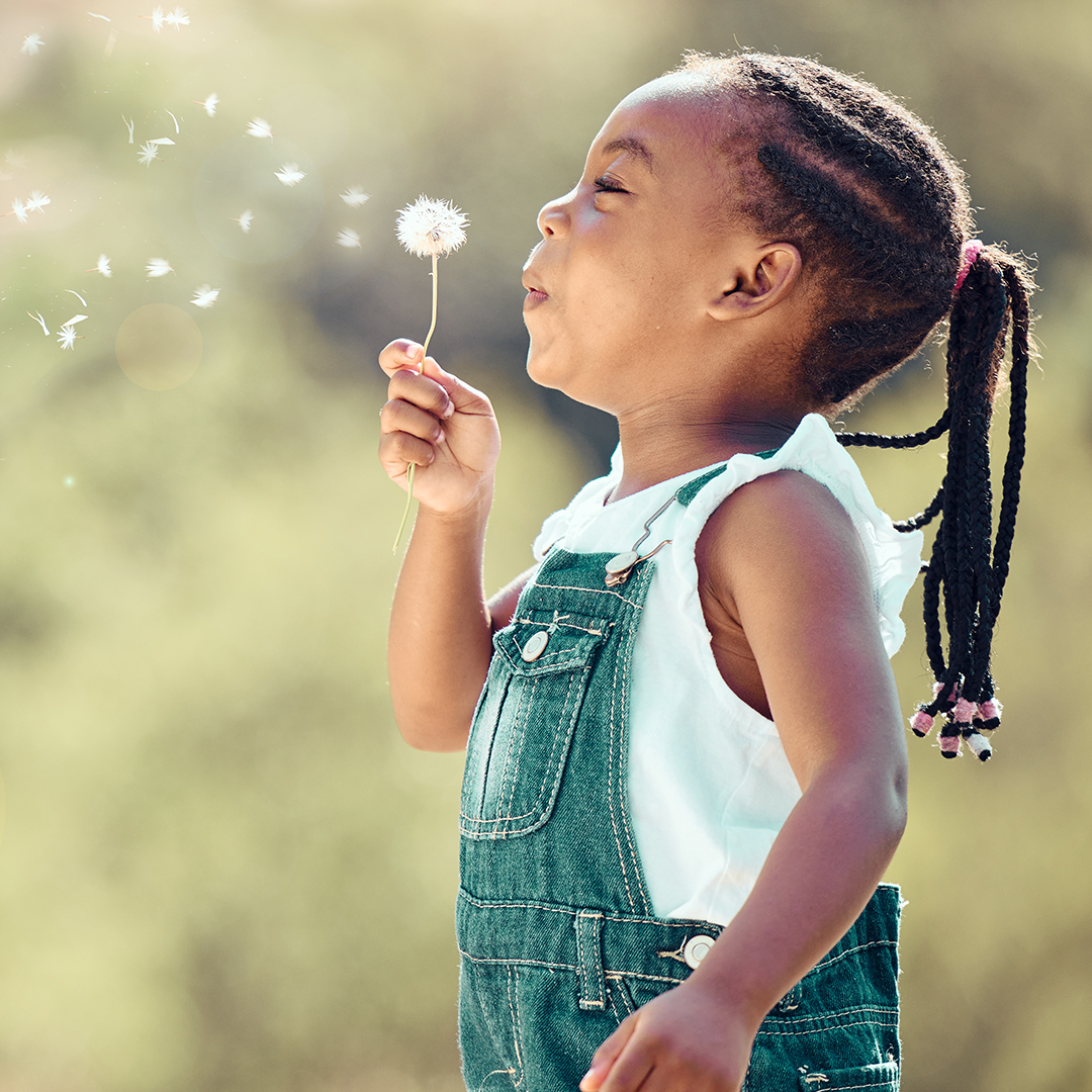 Little girl outside in overalls blowing on a flower