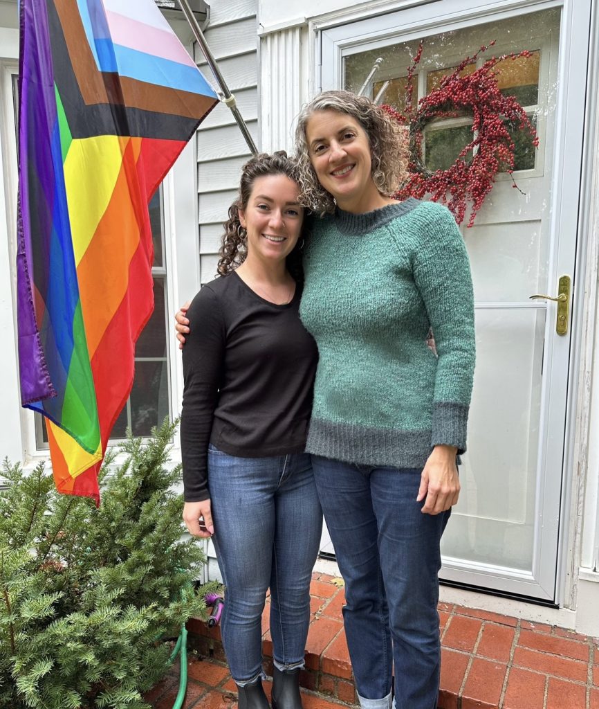Kris and Fiona standing outside of a home next to an LGBTQIA flag.