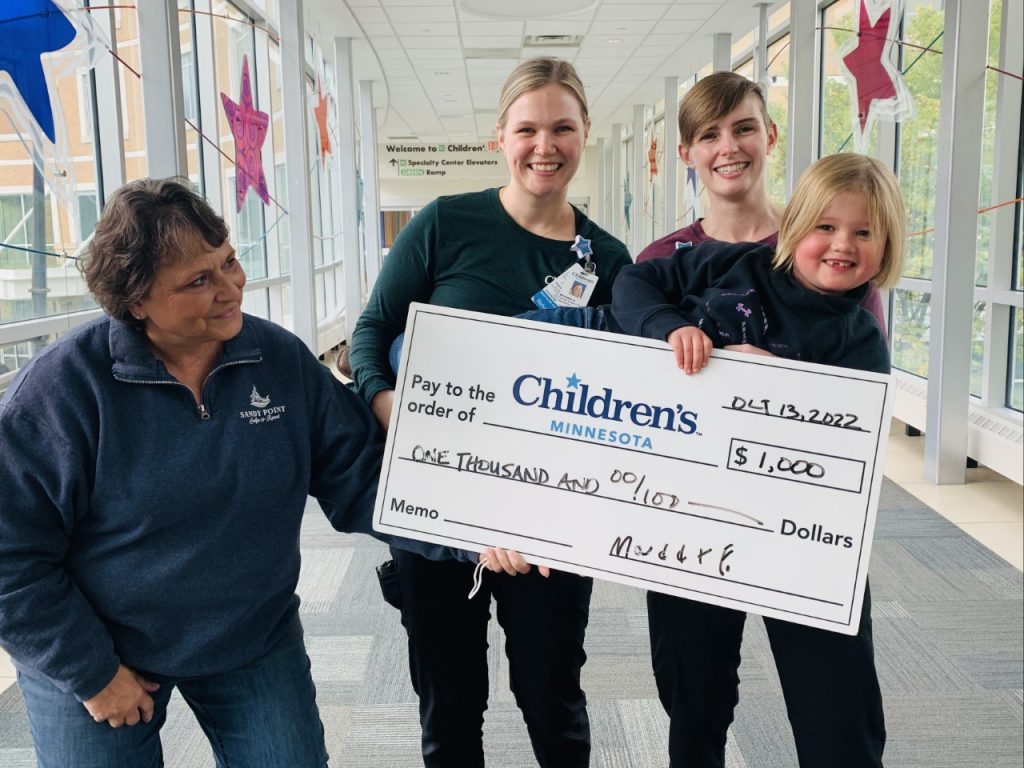 Three adults and a young girl hold up a novelty check made out to Children's Minnesota for $1,000. from Maddie