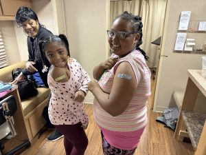 two young girls showing off their band aids after getting vaccinated to protect against the flu. Their guardian is in the background, smiling.