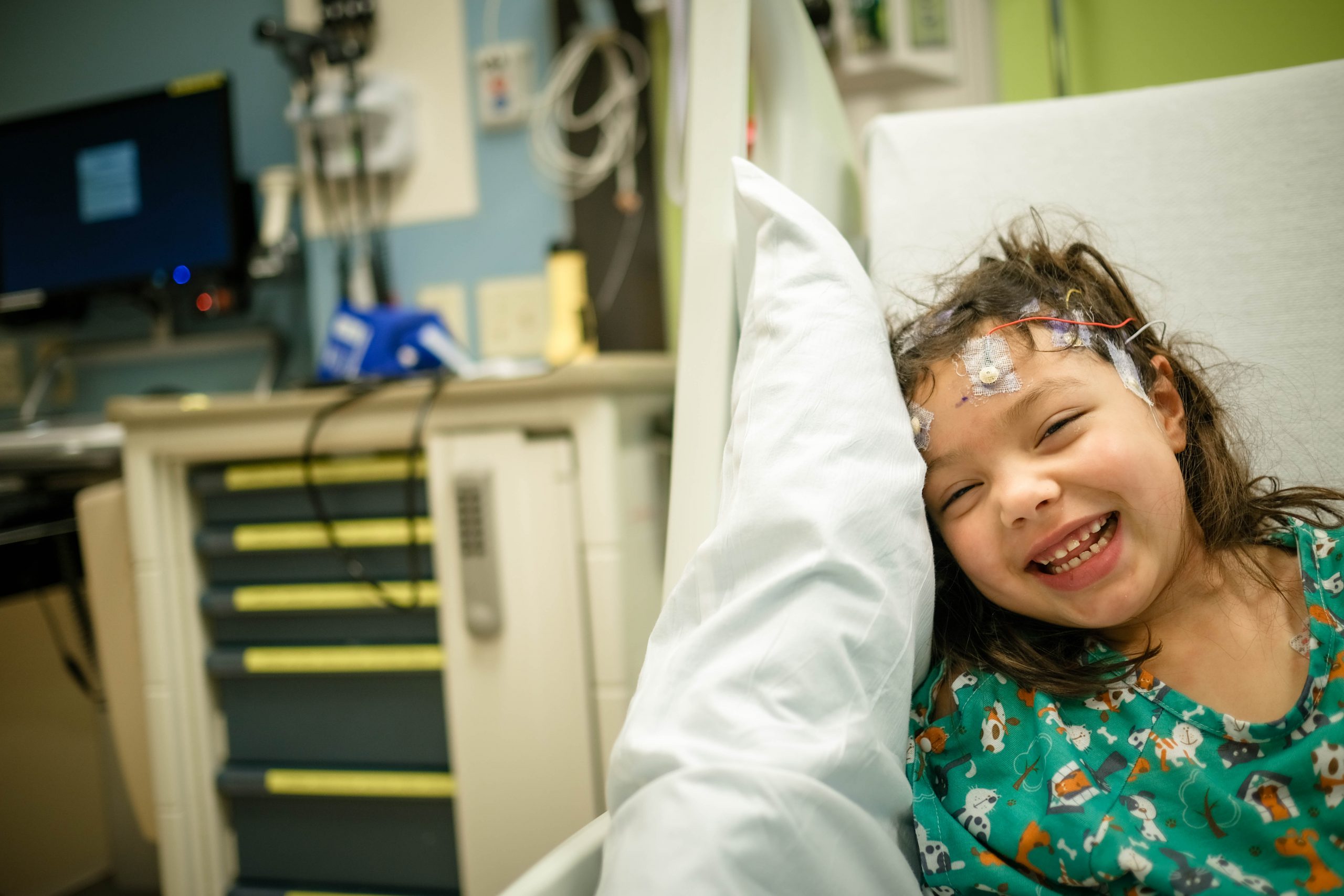 a child is in a hospital bed with medical wires connected to their forehead. They are in a hospital gown and smiling at the camera.