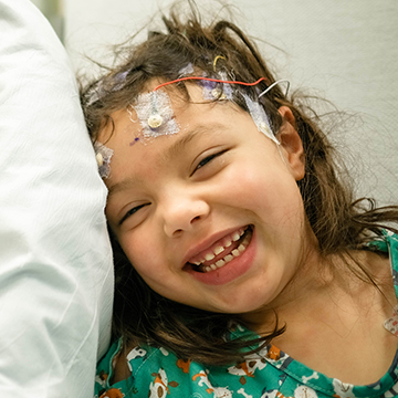 a child in a hospital bed smiles at the camera while wearing a hospital gown. She has wires taped to her forehead.