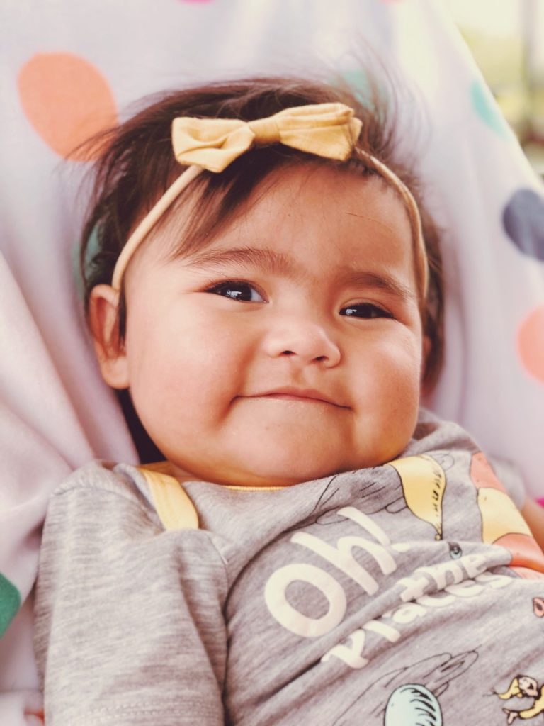 a young girl with a yellow bow in her hair is laying on a blanket. She has a small smile while looking at the camera.