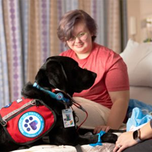 A teenage girl in a hospital bed is joined by a black dog, showing the healing environment of pet therapy.