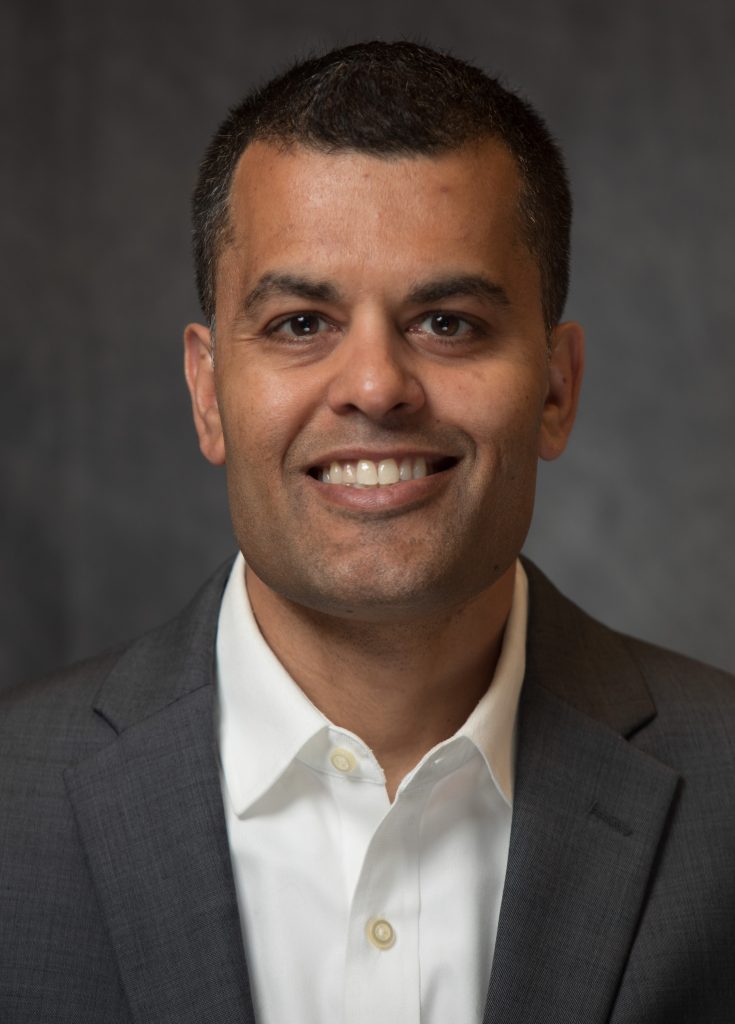 Sameer Gupta smiles, wearing a white collared shirt and gray suit jacket.