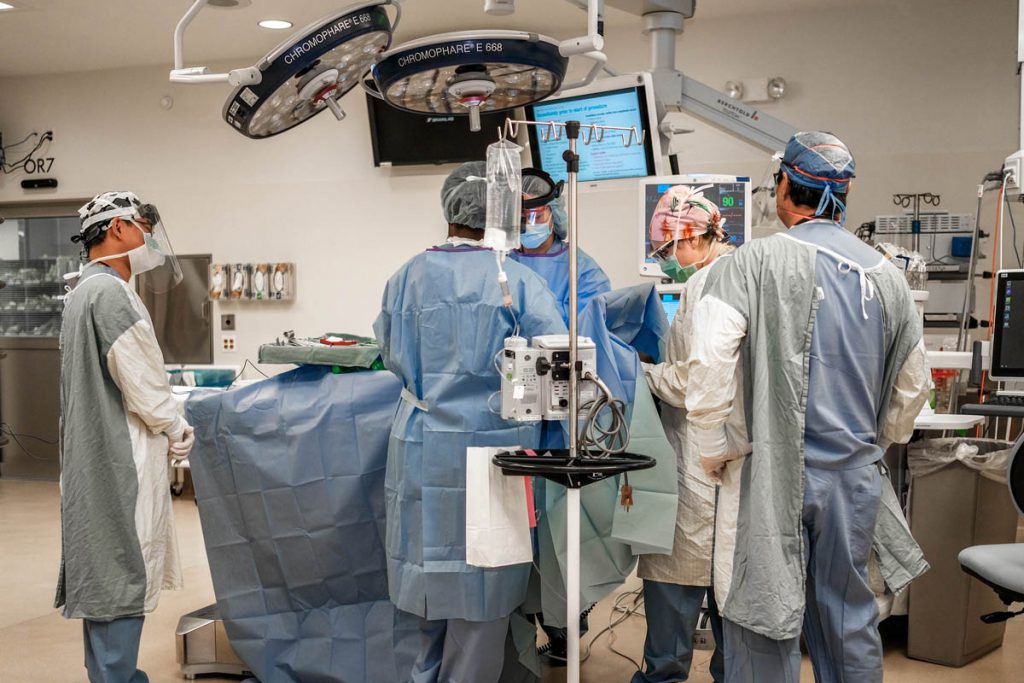 several surgeons are in full scrubs surround a patient on a bed in a surgery room.