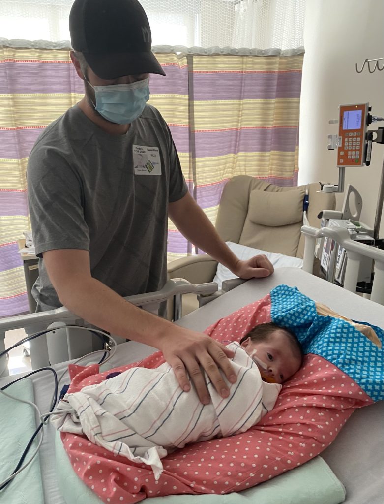 Noa in the hospital with a parent standing over her with a hand on her.