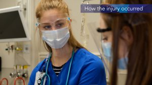 A woman with long blond hair pulled back, wears goggles, a mask and a face shield is speaking to someone who is blurred and partially off screen. On top of the image there is text that says "How the injury occurred."