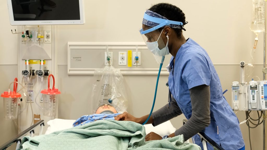 A caregiver wearing blue scrubs with a mask and face shield uses a stethoscope to examine a child on a hospital bed.
