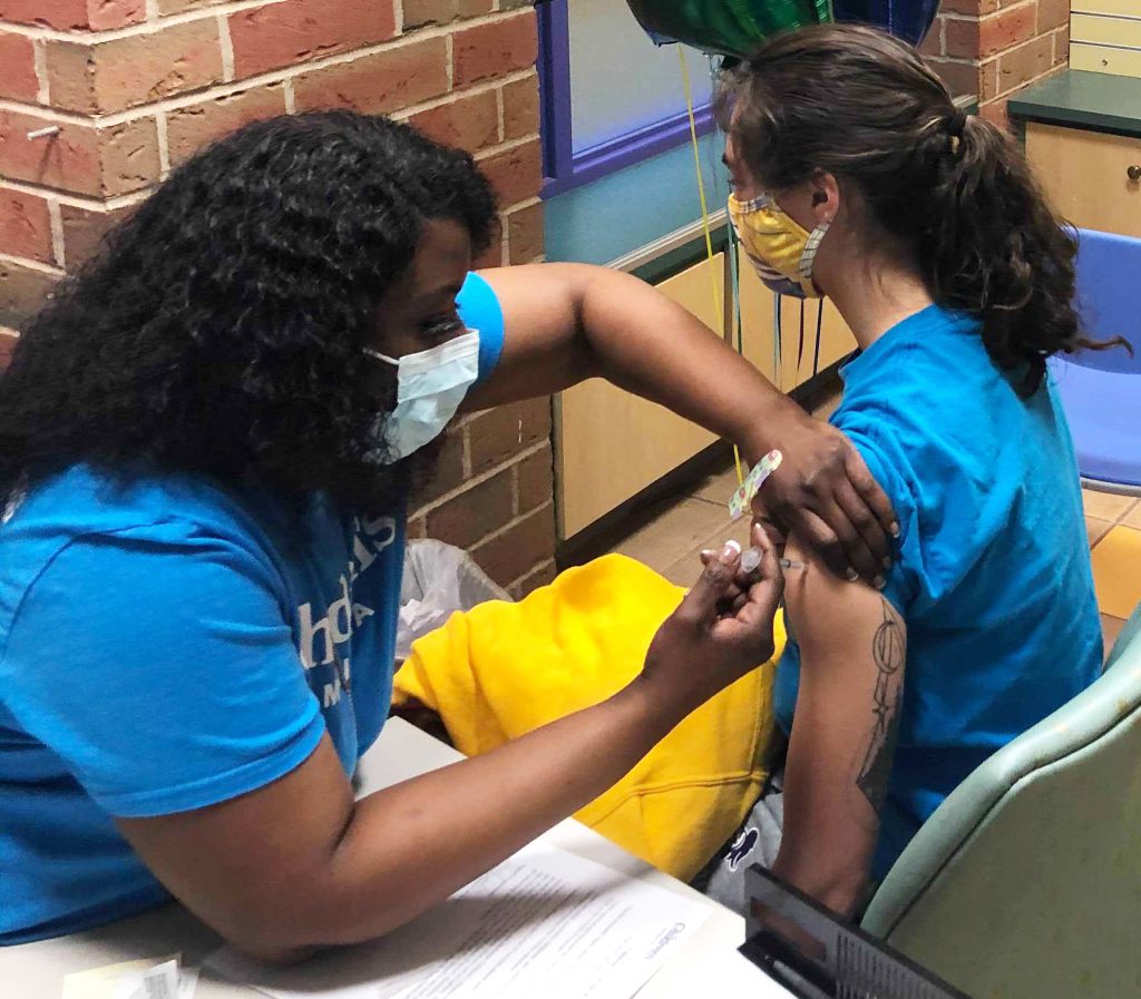 A Black woman with long curly dark hair, wears a mask and a blue t-shirt. She's administering a COVID-19 vaccine to a teenage girl with long dark hair and a blue t-shirt.