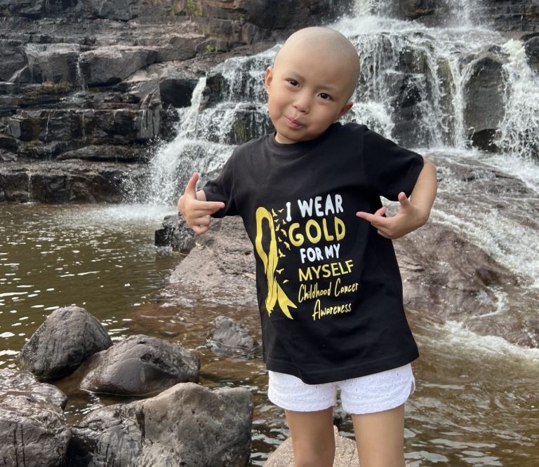 Valerie pointing at her shirt that reads 'I wear gold for myself. Childhood Cancer Awareness.'