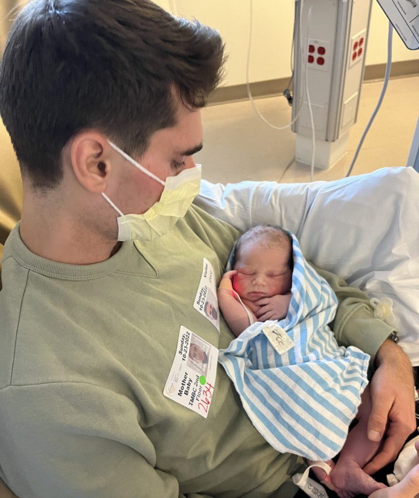 Joe in a facemask holding newborn William in the hospital