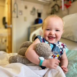 Xenia hugs a teddy bear while smiling at the camera. She's sitting in a hospital bed.