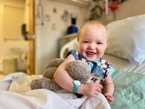 a toddler with short blonde hair holds a stuffed rabbit while sitting on a hospital bed. She is smiling at the camera.