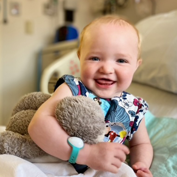 a toddler with short blonde hair holds a stuffed rabbit while sitting on a hospital bed. She is smiling at the camera.