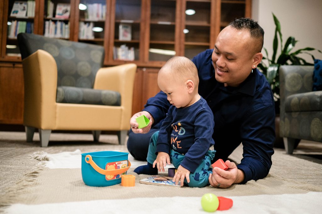 A father is sitting on the floor with his son. They are playing with toys.